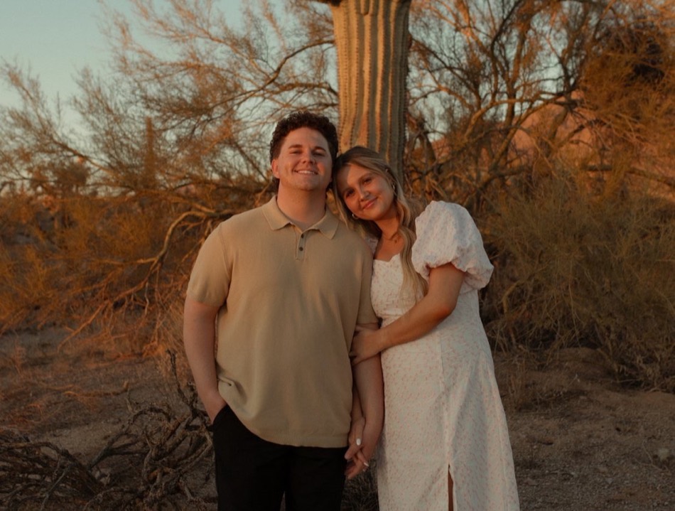 A smiling couple standing arm in arm in the Arizona desert at sunset, beneath a saguaro cactus
