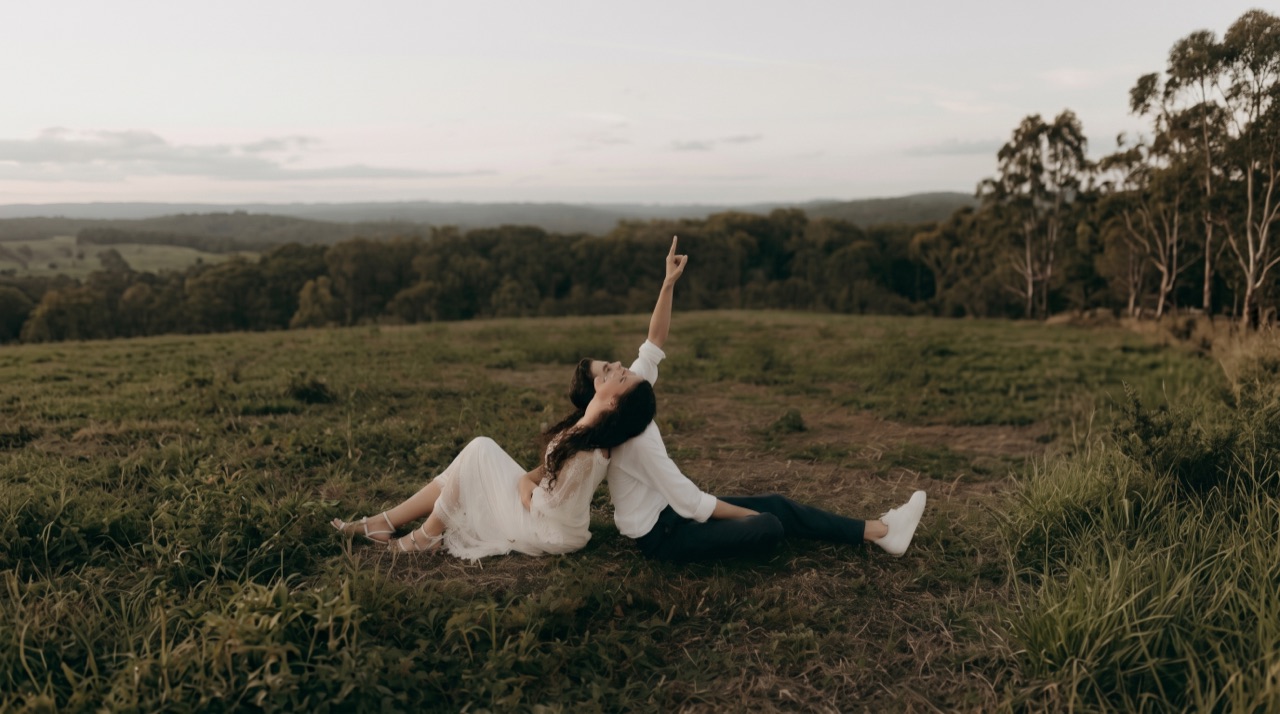 A couple sitting back to back in a hilltop field at dusk, one of them pointing at the sky